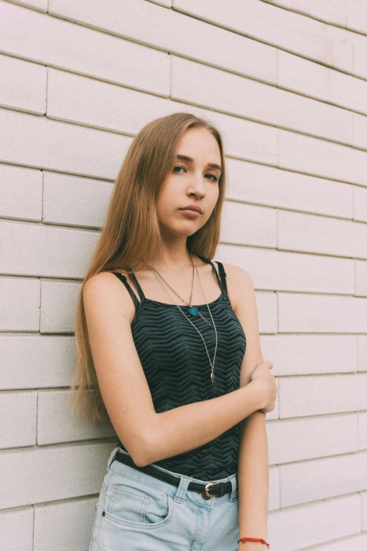 a young woman leaning against a white brick wall, inspired by Elsa Bleda, unsplash, neck chains, she is wearing a black tank top, young teen, amy sol in the style of