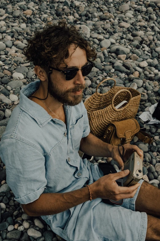 a man sitting on a rocky beach holding a cell phone, hozier, technological sunglasses, profile image, carrying a saddle bag