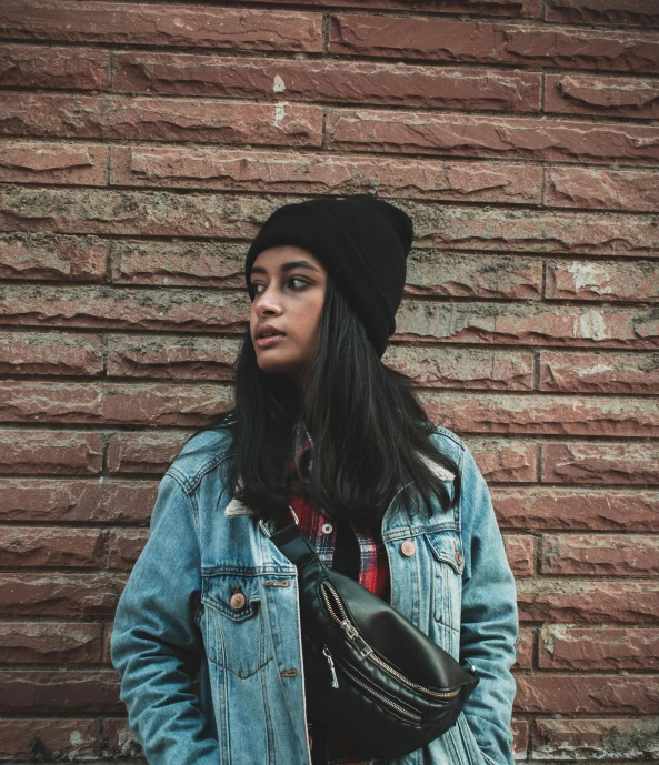 a woman standing in front of a brick wall, by Robbie Trevino, trending on pexels, beanie, young himalayan woman, jean jacket, student