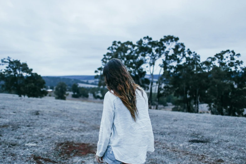 a woman standing in a field with trees in the background, by Lee Loughridge, trending on unsplash, happening, flowy hair standing on a rock, view from back, in the australian outback, blue and grey