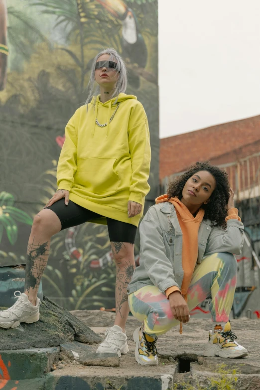 two people posing for a picture in front of a mural, a picture, inspired by Elsa Bleda, trending on pexels, graffiti, wearing a yellow hoodie, 2 techwear women, non binary model, pastel'