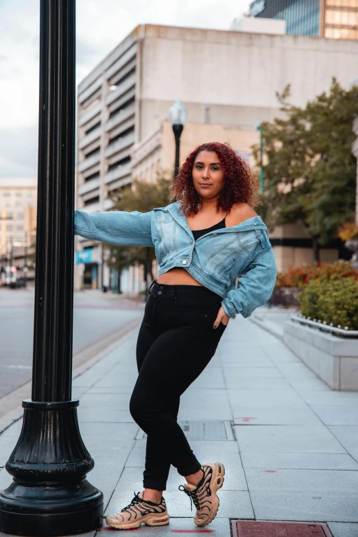 a woman leaning against a lamp post on a city street, an album cover, by Matt Cavotta, trending on pexels, realism, alluring plus sized model, wearing a jeans jackets, wearing a crop top, confident pose