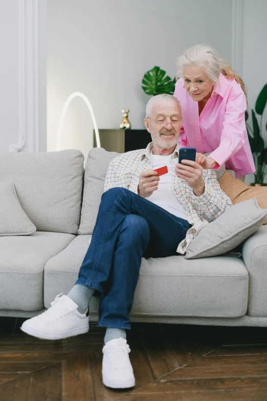 a man and woman sitting on a couch looking at a cell phone, gray haired, card game, inspect in inventory image, healthcare