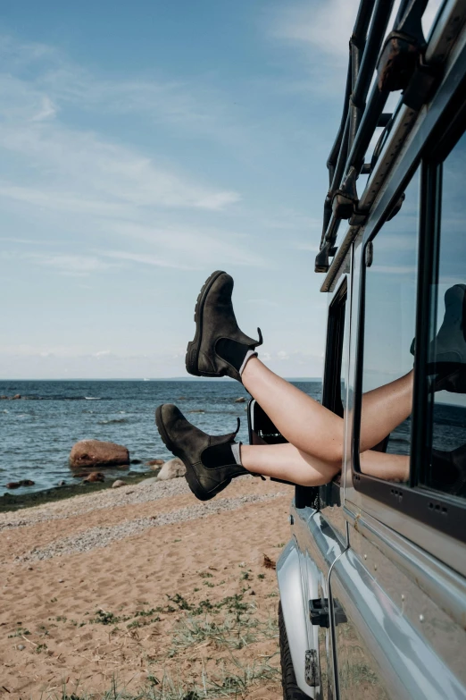 a woman's legs sticking out of a car window, by Julia Pishtar, trending on unsplash, is relaxing on a beach, doc marten boots, sitting on rocks, looking happy