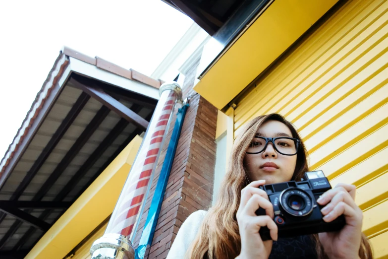 a woman taking a picture of herself with a camera, inspired by Vivian Maier, unsplash, photorealism, in front of ramen shop, wearing square glasses, on a yellow canva, fujifilm x100v