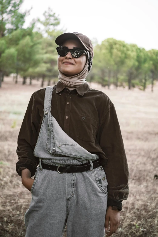 a man standing in a field with trees in the background, an album cover, by Ismail Acar, trending on unsplash, wearing overalls, hijab, with dark brown sunglasses, portrait androgynous girl