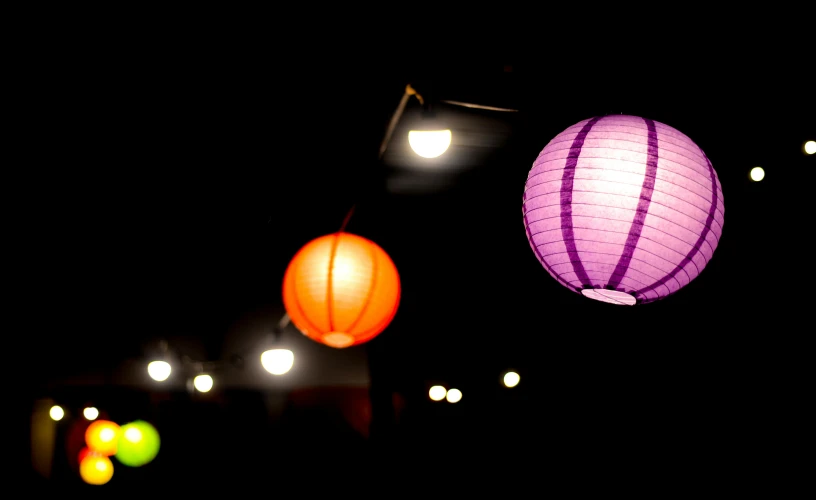 a couple of paper lanterns hanging from a string, by Matt Cavotta, pexels, light and space, deep purple and orange, rim lights purple and green, with a black dark background, cafe lighting