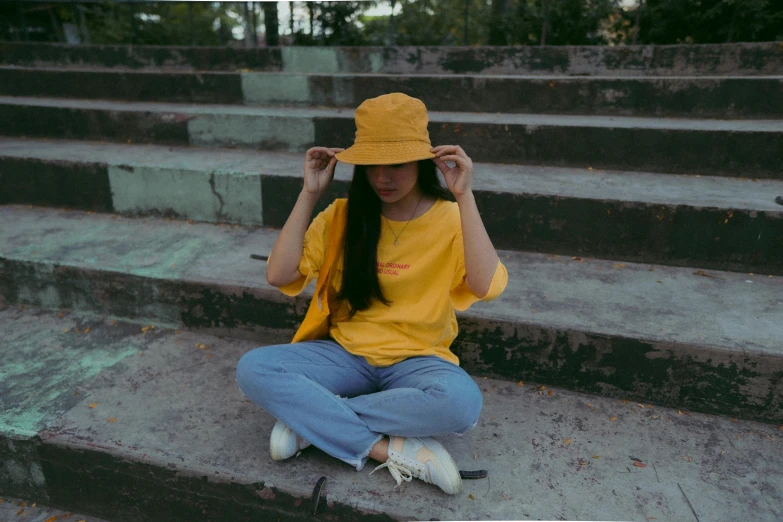 a woman sitting on steps talking on a cell phone, pexels contest winner, wearing a modern yellow tshirt, bucket hat, outlive streetwear collection, low quality photo