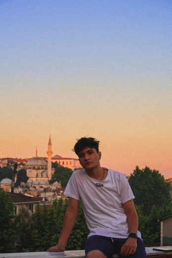 a man sitting on a ledge with a city in the background, an album cover, unsplash contest winner, istanbul, androgynous person, sunset glow around head, zayn malik