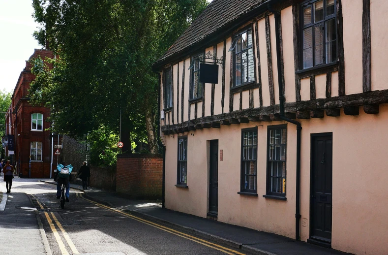 a couple of people riding bikes down a street, by IAN SPRIGGS, unsplash, arts and crafts movement, timbered house with bricks, the plague doctor, warwick saint, dusty street
