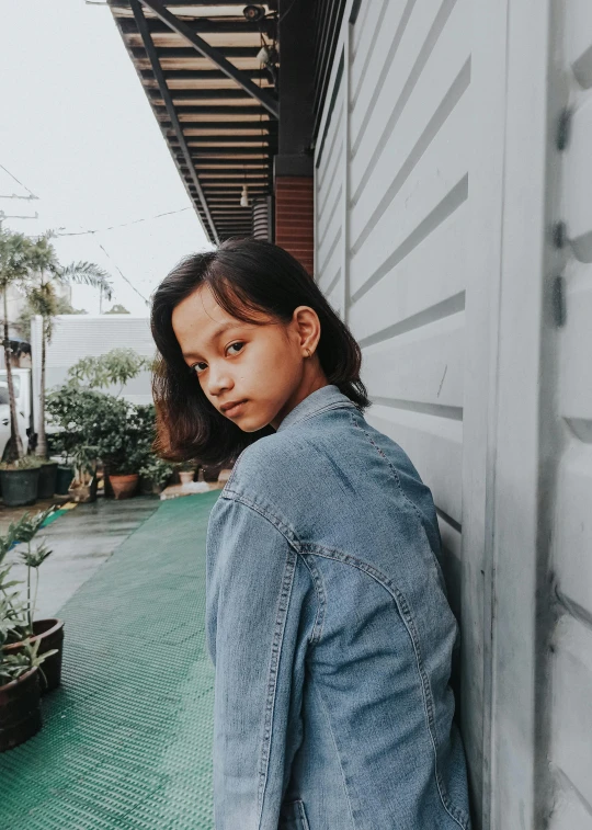 a woman leaning against a wall wearing a denim jacket, pexels contest winner, realism, south east asian with round face, boyish face, trending on vsco, album cover