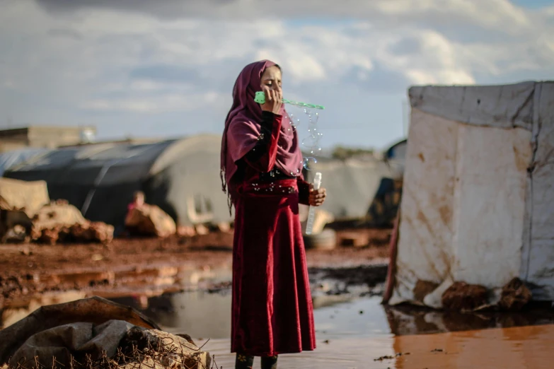 a woman standing next to a puddle of water, by Ibrahim Kodra, unsplash contest winner, hurufiyya, barrel fires and tents, drinking, refugees, soap bubbles