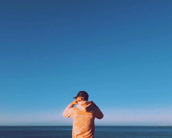 a man standing on a beach talking on a cell phone, unsplash, orange hoodie, cloudless blue sky, panoramic view of girl, mr beast