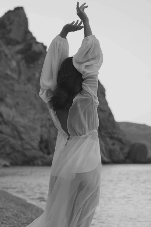 a woman standing on top of a beach next to a body of water, a black and white photo, by Alexis Grimou, romanticism, white sleeves, nico wearing a white dress, uploaded, faceless