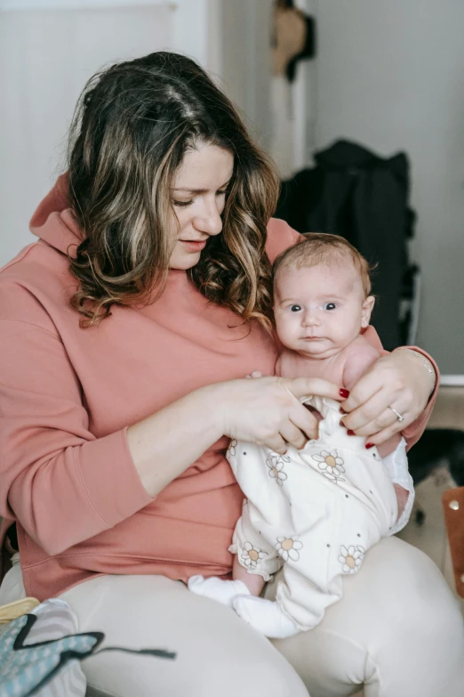 a woman holding a baby in her arms, by Alice Mason, pexels, wearing a pastel pink hoodie, sitting down, jen atkin, manuka