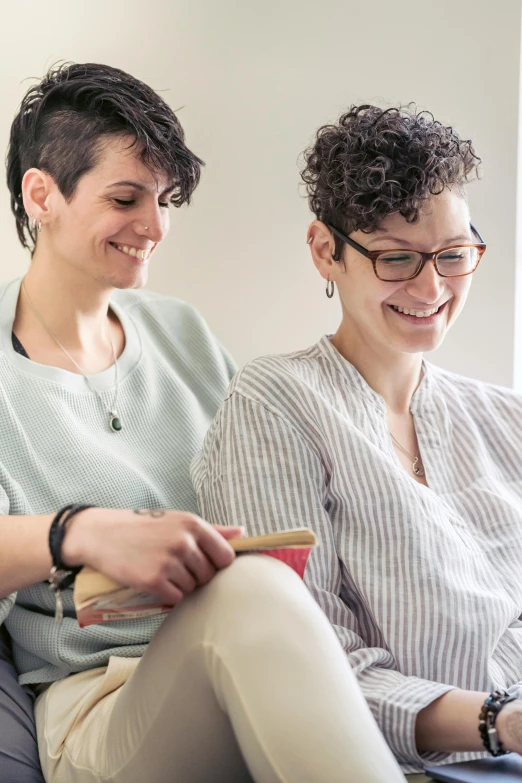 two women sitting on a couch looking at a laptop, by Harriet Zeitlin, non binary model, smiling couple, carefully crafted, promo image