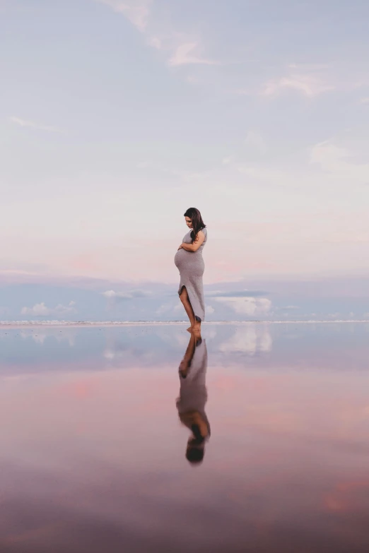 a woman standing on top of a large body of water, by Briana Mora, maternal photography 4 k, water reflection!!!!!, looking out at a pink ocean, leading to a beautiful