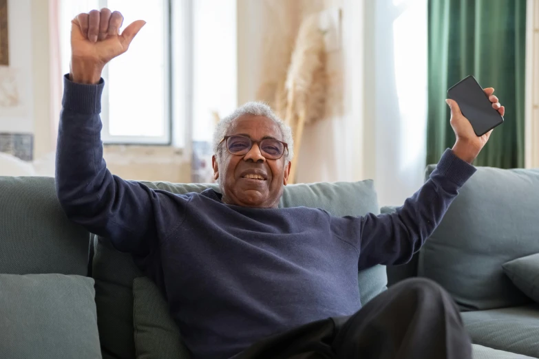 a man sitting on a couch holding up a cell phone, inspired by Ismail Gulgee, pexels contest winner, arms stretched out, he is about 8 0 years old, arms raised, at home