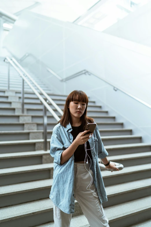 a woman standing in front of a set of stairs, by Jang Seung-eop, trending on pexels, happening, earbuds, she is holding a smartphone, young asian girl, grey