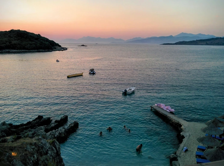 a group of people swimming in a body of water, by Alexis Grimou, pexels contest winner, which shows a beach at sunset, greek setting, pink white turquoise, mountains and ocean