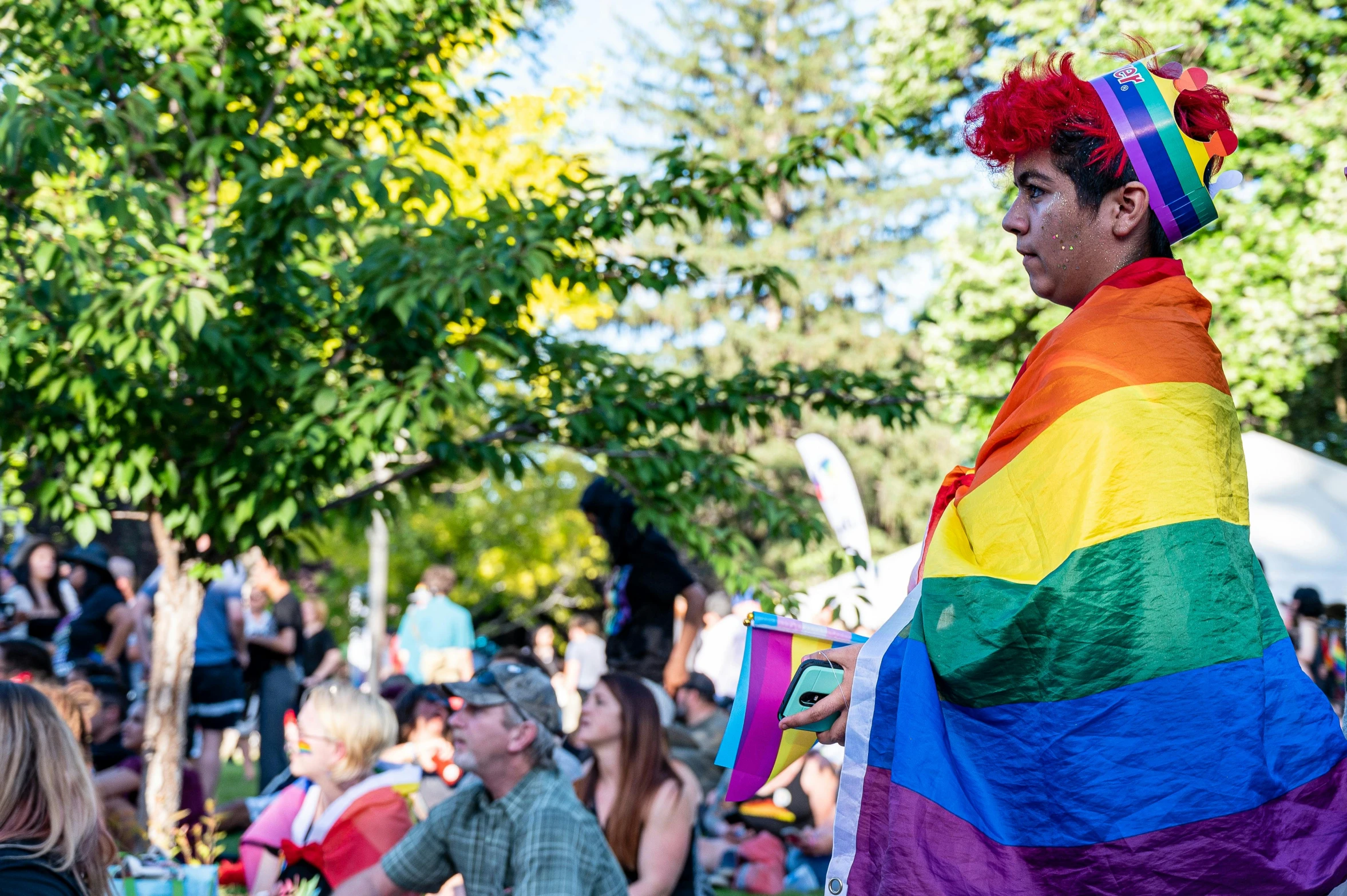 a woman standing in front of a crowd holding a rainbow flag, process art, city park, costume, looking to his side, square