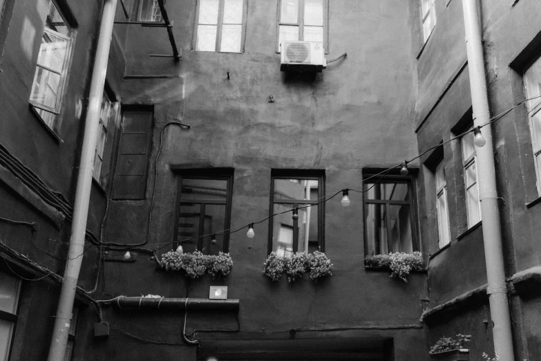 a black and white photo of an alleyway, a black and white photo, wires hanging across windows, lamps and flowers, nikolay kopeykin, low quality photo