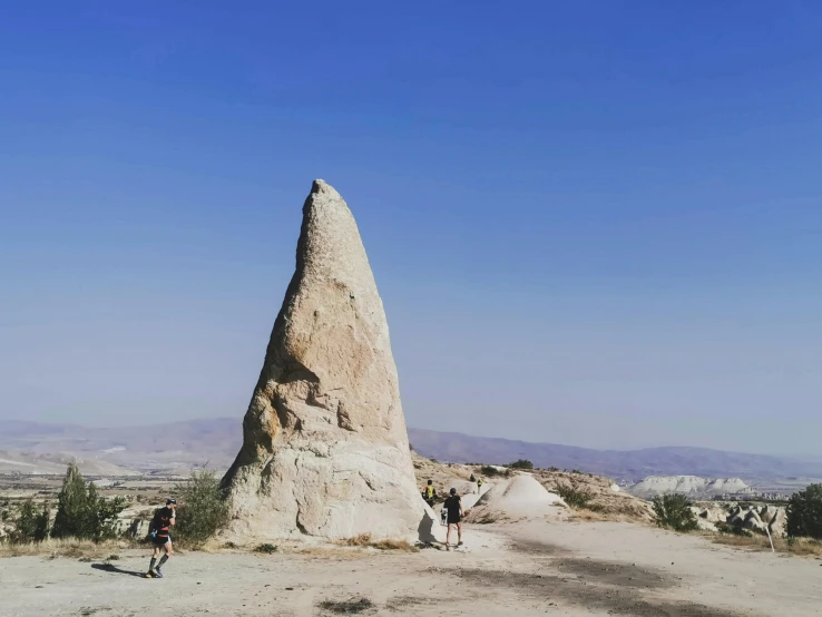 a man riding a bike down a dirt road next to a tall rock, a marble sculpture, by Muggur, people walking around, spire, background image, turkey