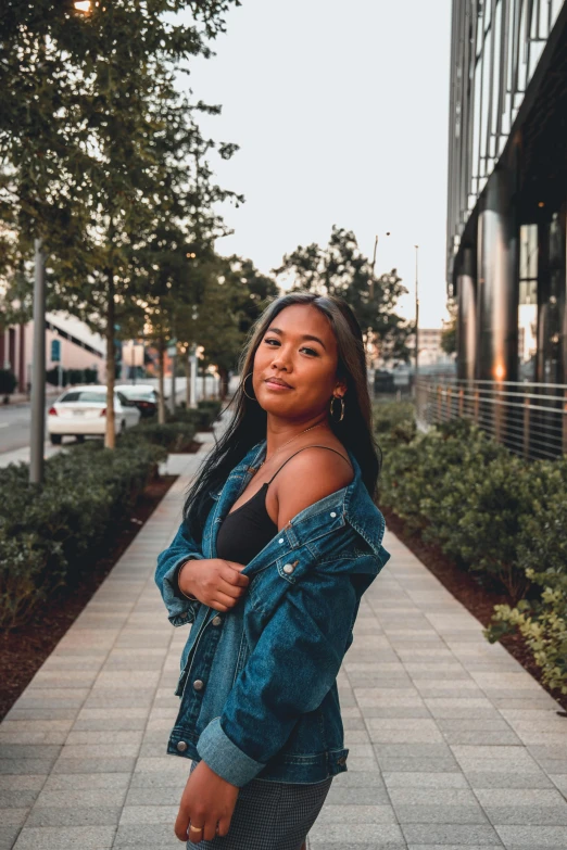 a woman standing on a sidewalk in front of a building, by Robbie Trevino, wearing a jeans jackets, a young asian woman, portrait featured on unsplash, african american young woman