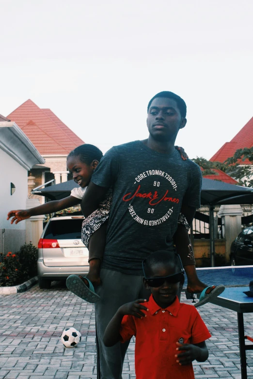 a man standing next to a little boy with a soccer ball, an album cover, by Chinwe Chukwuogo-Roy, pexels contest winner, happening, in front of the house, wearing a t-shirt, rides, ceo