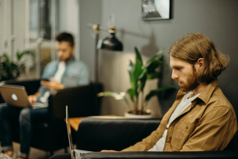 a man sitting on a couch using a laptop, trending on pexels, medium shot of two characters, sitting behind desk, sideways glance, no watermarks