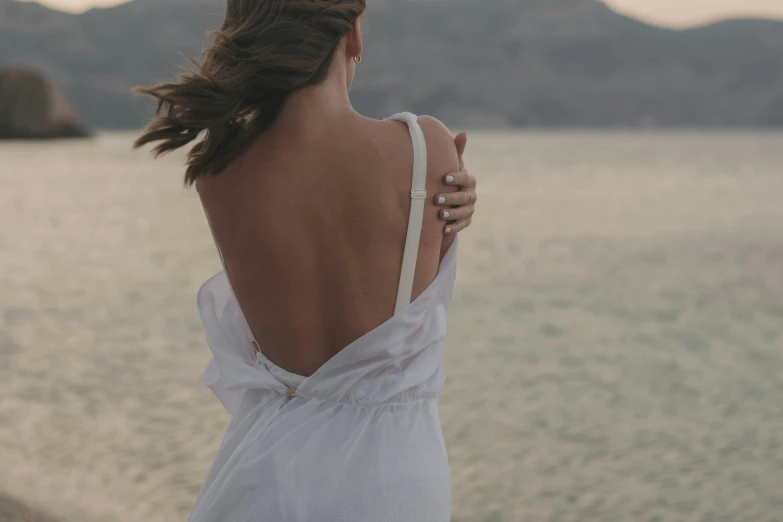 a woman in a white dress standing on a beach, pexels contest winner, romanticism, showing her shoulder from back, early evening, photoshoot for skincare brand, soft silk dress