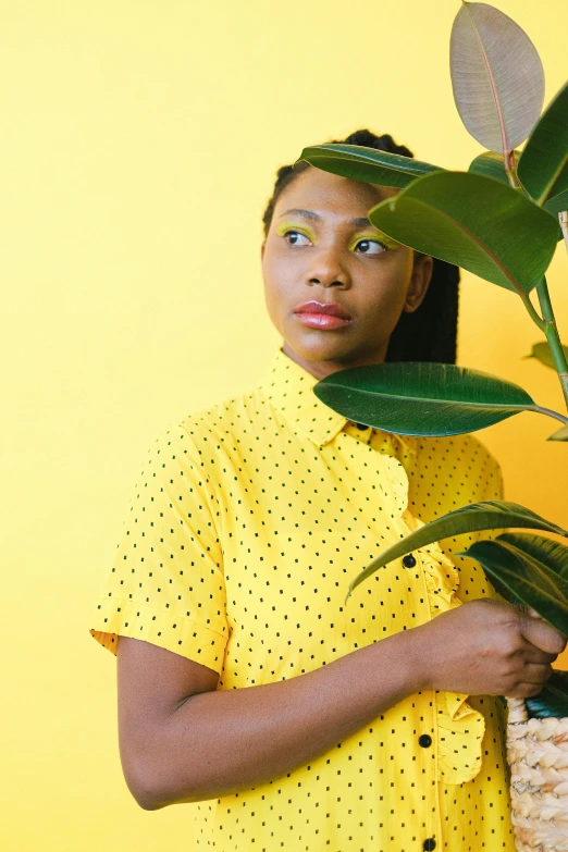 a woman holding a potted plant in front of a yellow wall, by Dulah Marie Evans, green shirt, next to a tree, promo image, black young woman