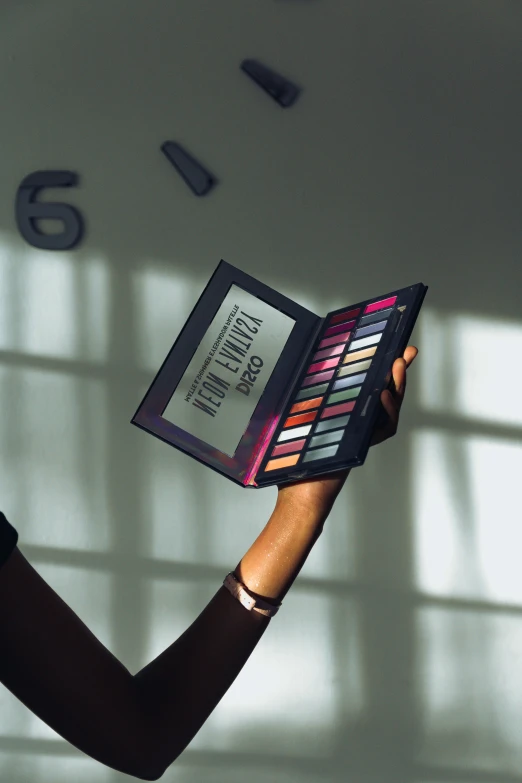 a woman holding a makeup palette in front of a clock, happening, dark skinned, iridescent palette, matte finish, revolutionary