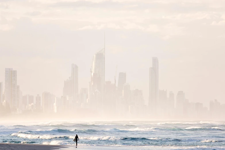 a man standing on top of a beach next to the ocean, by Peter Churcher, unsplash contest winner, minimalism, city skyline, morning haze, gold coast australia, viewed from the ocean