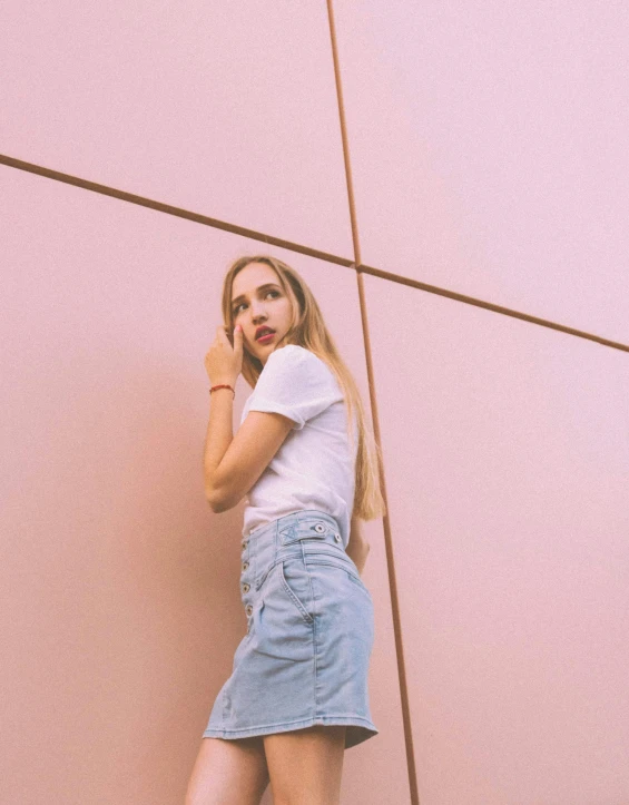 a woman standing in front of a wall talking on a cell phone, an album cover, inspired by Elsa Bleda, trending on pexels, aestheticism, pink skirt, jeans and t shirt, profile image, sydney sweeney