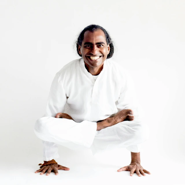 a man sitting on top of a white floor, anjali mudra, dramatic smiling pose, hugging his knees, front and center