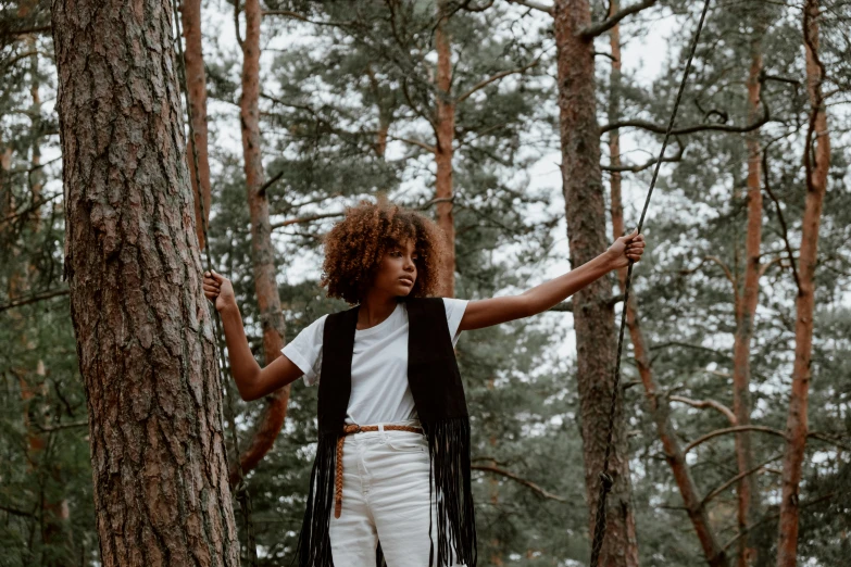 a woman standing next to a tree in a forest, by Emma Andijewska, pexels contest winner, afrofuturism, outfit : jeans and white vest, black teenage girl, long trunk holding a wand, concert