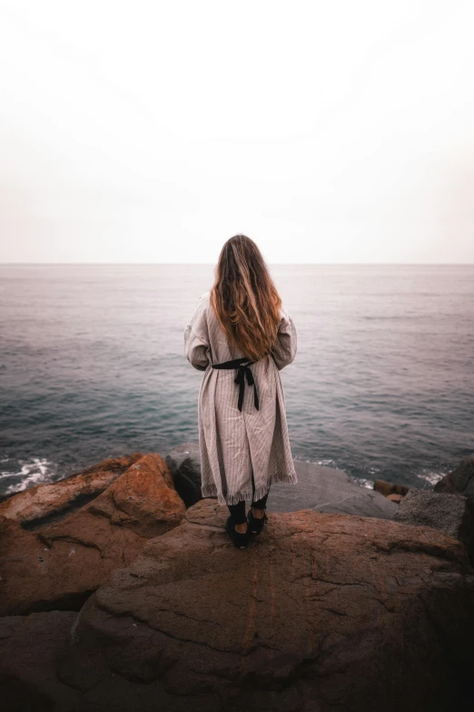 a woman standing on top of a rock next to the ocean, unsplash, romanticism, dressed in a robe, facing away from camera, teenage girl, grey