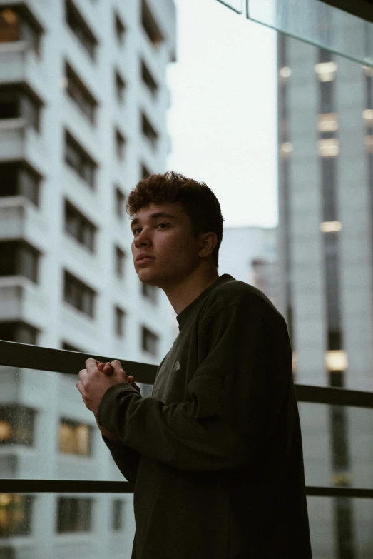 a man standing on top of a balcony next to a tall building, an album cover, inspired by John Luke, pexels contest winner, looking away from camera, portrait soft low light, boyish face, ansel ]