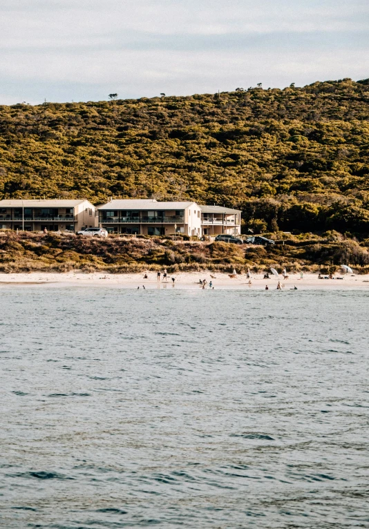 a group of people riding surfboards on top of a body of water, several cottages, australian beach, white sand beach, seen from a distance