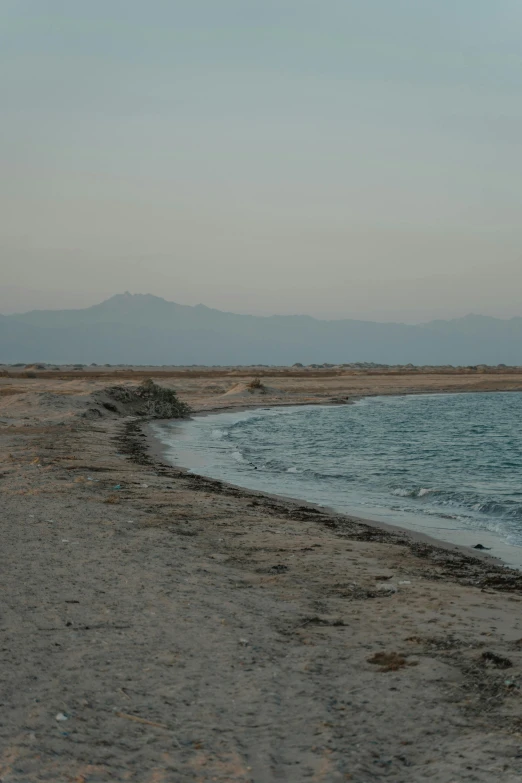 a man standing on top of a sandy beach next to the ocean, les nabis, al - qadim, distant mountains, low light, tar - like