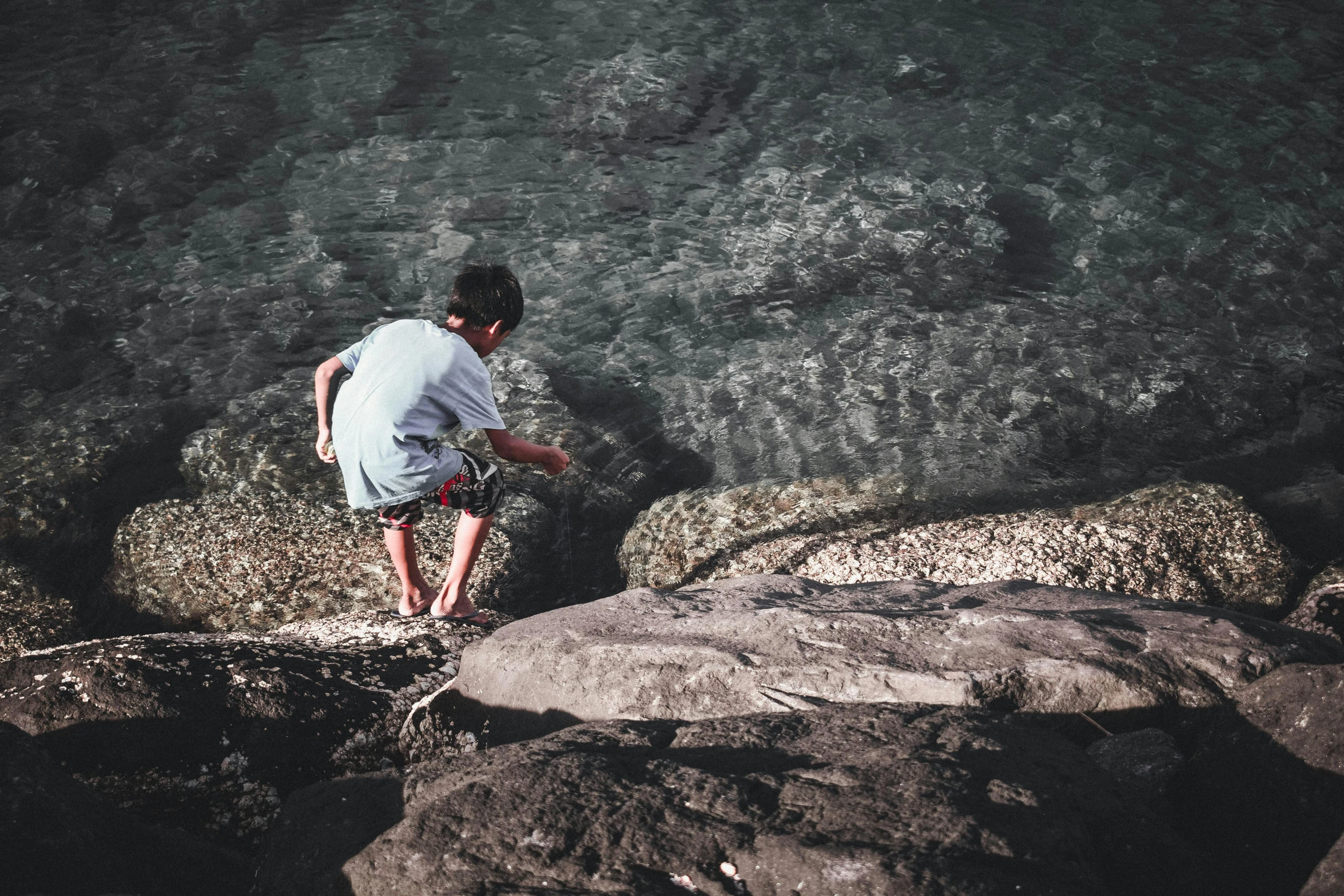 a man standing on top of a rock next to the ocean, pexels contest winner, happening, walking boy, playing with the water, rocky roads, crouching