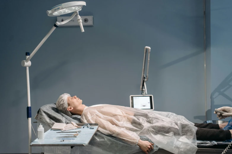 a person laying on a bed in a room, by Évariste Vital Luminais, pexels contest winner, massurrealism, surgical gown and scrubs on, cyber augmentation implant, angle view, on an operating table