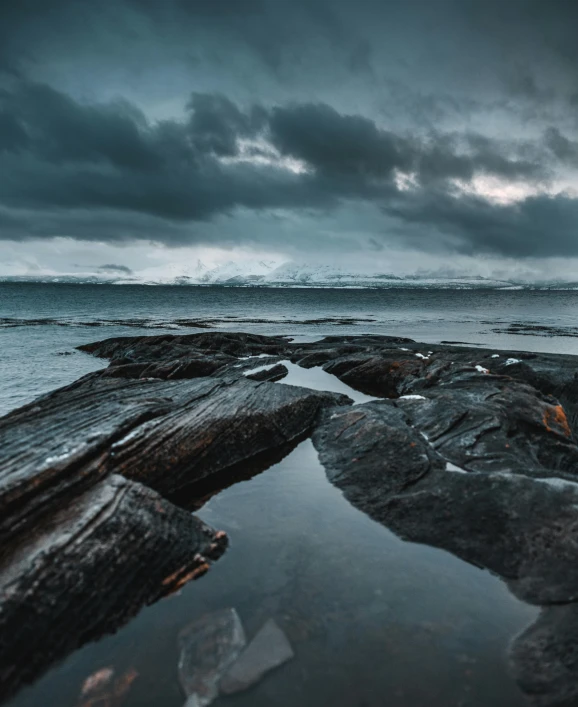 a large body of water sitting on top of a rocky beach, by Jesper Knudsen, dark clouds in the distance, high-quality photo, trending photo, jagged blocks of stone