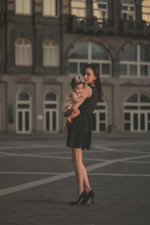 a woman in a black dress holding a baby, by Fei Danxu, pexels contest winner, in a city square, cute elegant pose, dim lit, 15081959 21121991 01012000 4k