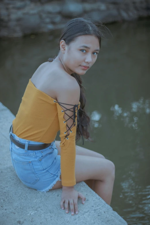 a woman sitting on a ledge next to a body of water, by Reuben Tam, pexels contest winner, realism, wearing yellow croptop, halfbody headshot, long sleeves, wearing a camisole and shorts