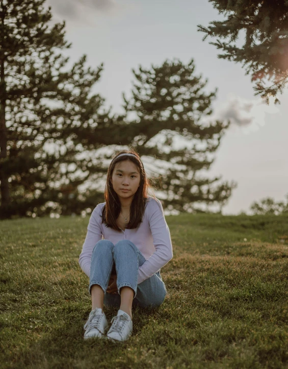 a woman sitting on top of a lush green field, wearing a pastel pink hoodie, leslie zhang, wearing white sneakers, low lighting