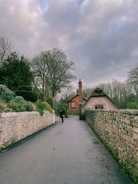 a person walking down a road next to a stone wall, inspired by Stanley Spencer, unsplash, small cottage in the foreground, gloomy skies, at highgate cemetery, winter setting