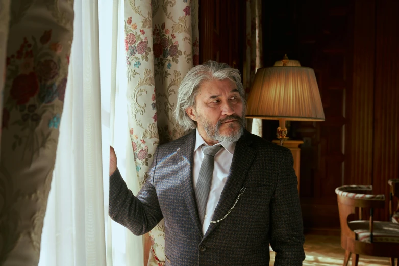 a man in a suit standing in front of a window, inspired by Alexander Roslin, trending on pexels, baroque, gray hair and beard, movie scene, fully covered in drapes, dressed in 1970s menswear