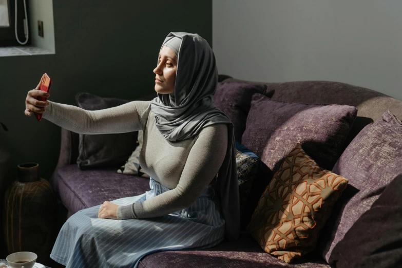 a woman sitting on top of a purple couch, a portrait, inspired by Maryam Hashemi, unsplash contest winner, hurufiyya, wearing a grey robe, concerned expression, hijab, at home